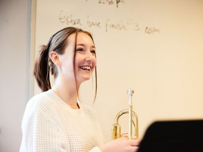A girl in a white jumper is holding a trumpet. She has a big smile on her face